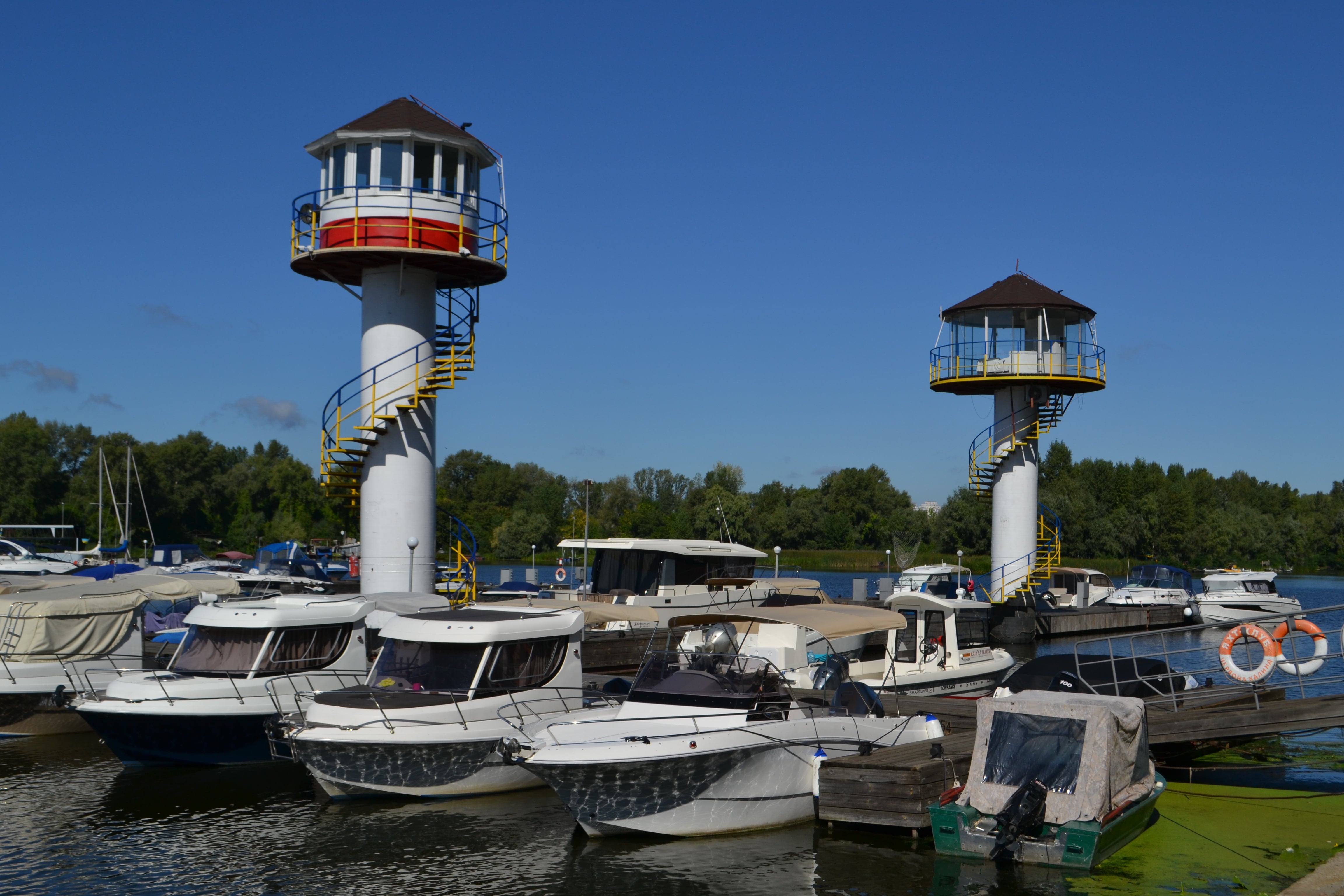 Яхт-клуб Червона Калина – два невеликих маяка над водою із заскленими приміщеннями в горі. Катери та яхти припарковані уздовж пірса на річці Дніпро.  